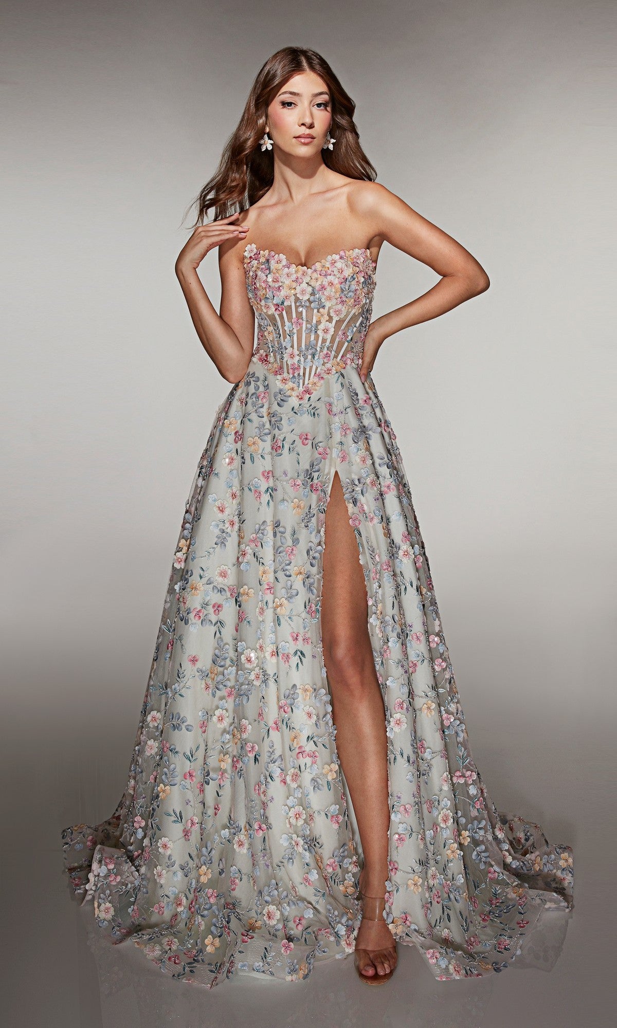 Wearing the Alyce 61986 Long Prom Dress, a woman poses against a plain background. The strapless gown features a fitted bodice, 3D floral appliques, pastel embroidered flowers, a high slit, and an elegant floor-length silhouette.